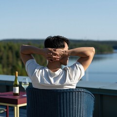 Obraz premium Young man leaning back on a terrace chair overlooking calm lake with wine for summer wallpaper and travel background design