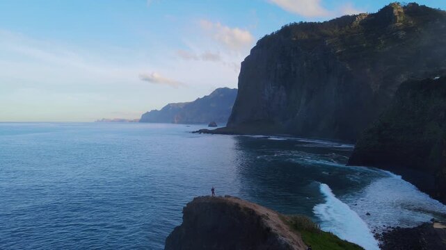 A lone person walks along dramatic coastal cliffs overlooking the ocean and towering mountains, creating a cinematic adventure scene with natural sea and wind sounds.