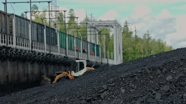Tracking shot excavator loading coal mound at industrial dock of train freight railway car near elevated conveyor walkway.