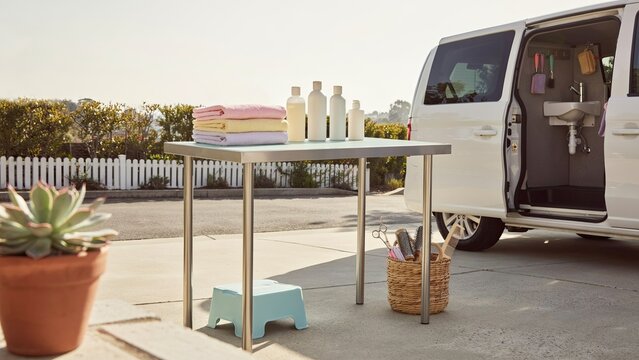 Steel table holds stacked linens near a parked van containing a portable sink in an outdoor residential driveway setting.