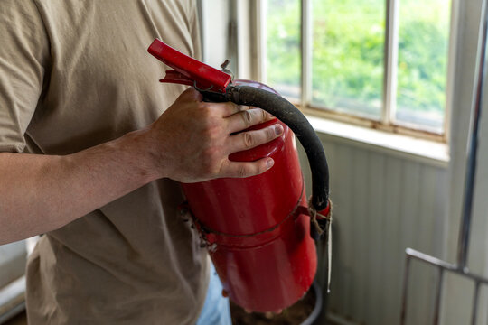 Man holding a red portable fire extinguisher in hands indoors near a window, preparing for emergency response and safety management.
