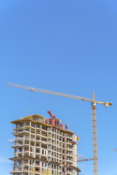 A tall yellow tower crane operating at a high-rise residential building construction site. A multi-story concrete framework of the developing skyscraper with scaffolding and insulation layers.
