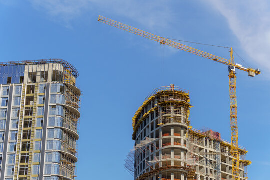 A tall yellow tower crane operating at a high-rise residential building construction site. A multi-story concrete framework of the developing skyscraper with scaffolding and insulation layers.