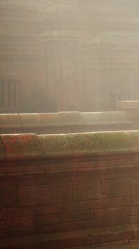 Sunlight filters through mist in a historical area, illuminating an old brick wall. The setting shows ancient structure hints and soft light in the early hours of the day.