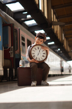 Young boy sitting on vintage suitcase holding large clock at station