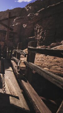 A wooden path runs alongside rocky cliffs in a natural area. Sunlight shines down, casting shadows on the ground. The scene shows rough textures of rocks and wood.