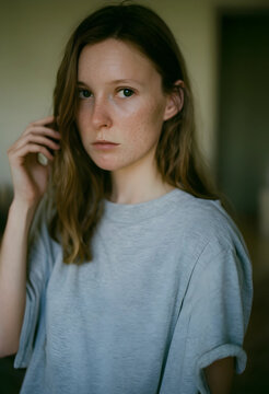 Young woman with freckles and long brown hair wearing a gray t-shirt, looking directly at the camera with a neutral expression