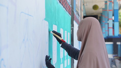 Female ArtistWearing a Hijab Painting a Large Outdoor Mural with Turquoise Background © chandra
