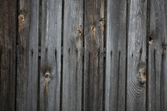 Close-up of a weathered wooden wall with vertical planks. Features a textured surface of dark brown and gray timber, organic wood knots, and a central horizontal row of rusted metal nails.
