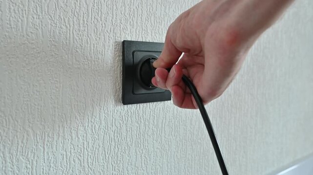 Close up view of a person's hand inserting a black power plug into a matching black electrical socket on a textured white wall, symbolizing the concepts of connection and electricity supply