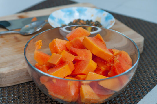 Glass bowl filled with fresh papaya cubes beside a cutting board, knife and removed seeds. Tropical fruit prepared for healthy eating and homemade recipes.