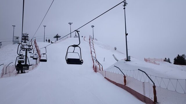 There are chairlifts on the slopes of the ski center. Skiers climb them. There are ski trails in the snow and ski trails are visible. For safety there is a mesh fence and metal lampposts. Overcast