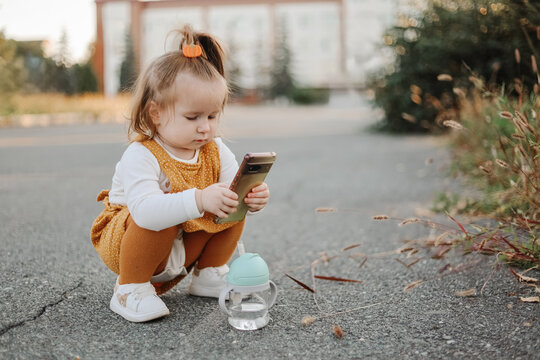 Toddler using smartphone outdoors on pavement near sippy cup