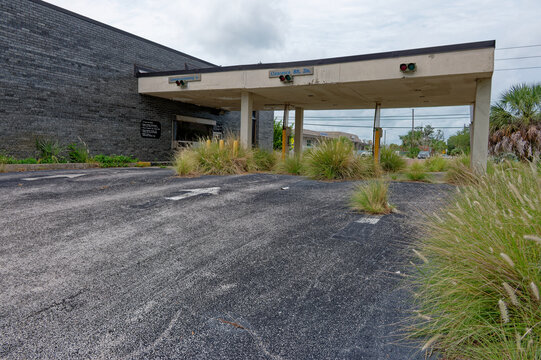 An abandoned bank building with a cracked canopy and overgrown asphalt drive beneath a cloudy sky, surrounded by palms and weeds, evoking decay, neglect and eerie suburban desolation.
