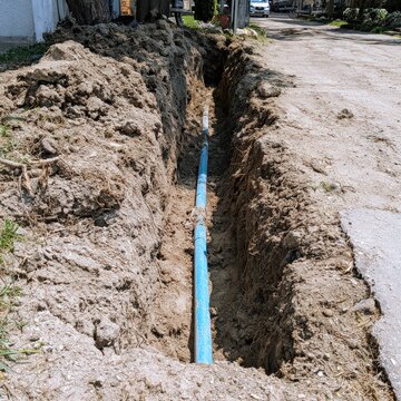 Narrow excavation beside driveway with blue service pipe in midday sun