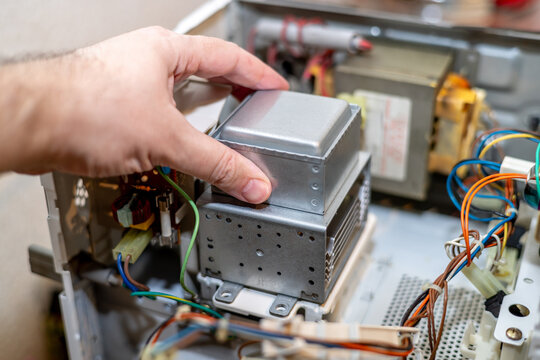 A technician's hand holds a microwave oven magnetron during repair. Colored wires, connectors, and adjacent service parts are visible