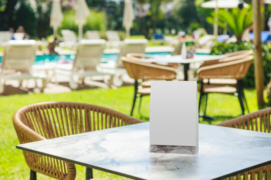 Blank vertical table tent mockup placed on a marble table beside an outdoor swimming pool at a leisure resort. The empty display provides clean copy space for restaurant menus, promotional offers