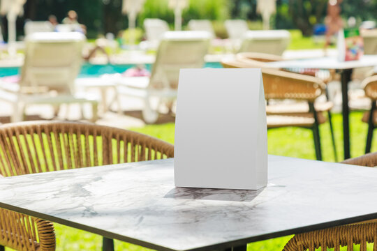 Blank vertical table tent mockup placed on a marble table beside an outdoor swimming pool at a leisure resort. The empty display provides clean copy space for restaurant menus, promotional offers