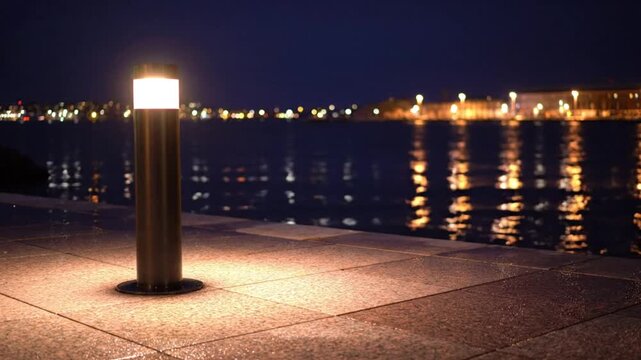 Bright bollard light illuminates waterside promenade at night with city lights reflecting in water