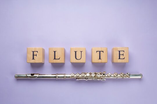 Silver flute instrument placed next to wooden letter blocks spelling flute on a light purple background
