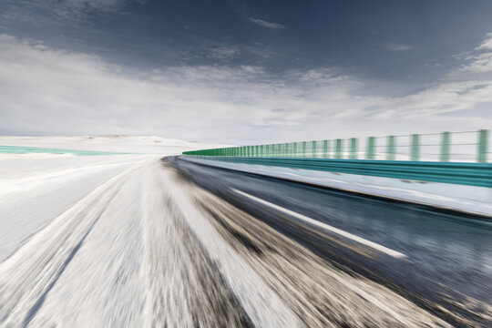 A snow-covered road with motion blur stretches alongside a green guardrail under an overcast sky, showing a wintry outdoor transport scene.