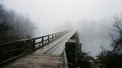 Fototapeta premium Foggy morning scene with wooden bridge extending into dense mist over river.