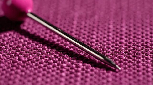 Close-up of a sewing pin with a pink head resting on textured violet fabric