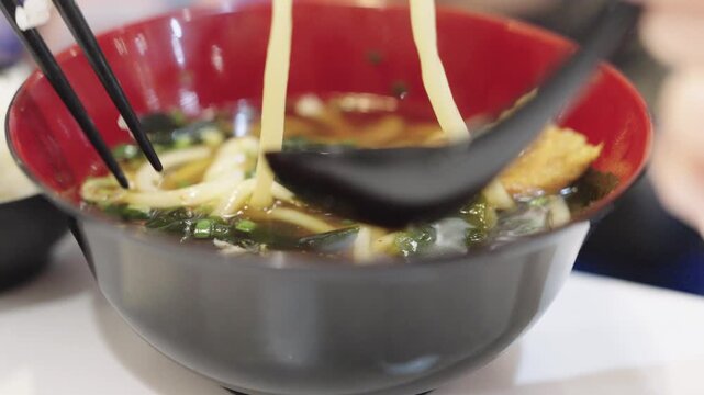 Close-up of chopsticks lifting thick udon noodles and seaweed from a bowl of hot Japanese soup. Professional food photography highlighting traditional Asian cuisine and authentic dining experience.