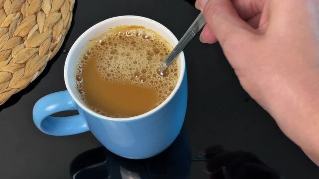Close-up of a hand thoroughly mixing instant coffee with condensed milk and hot water using a teaspoon until smooth.