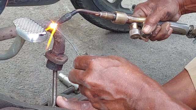 A close-up view of a skilled welder using a gas torch and metal tool or filler rod to heat and solder a leaking motorcycle exhaust pipe during an outdoor repair process.