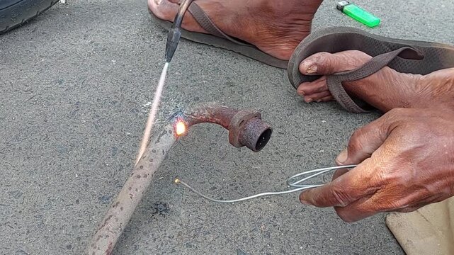 A close-up view of a skilled welder using a gas torch and metal tool or filler rod to heat and solder a leaking motorcycle exhaust pipe during an outdoor repair process.