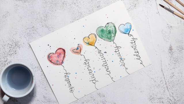 Hand writes lettering on handmade watercolor greeting card decorated with colorful heart illustrations. Top view shows creative drawing process on textured paper with brush pens, paintbrushes and cup