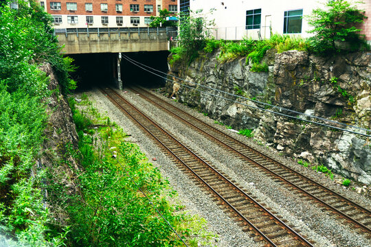 An industrial railway scene showing dual tracks leading into a deep dark tunnel set amidst rocky walls, lush green vegetation, and an urban building exterior.
