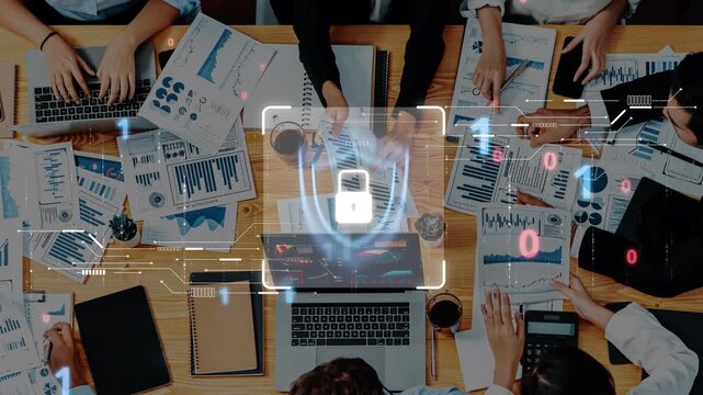 A birds-eye view of a team engaged in a collaborative meeting surrounded by laptops and various documents displaying data analysis metrics and security. Raster
