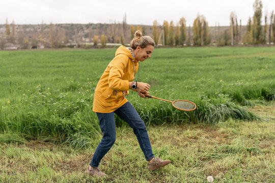 Woman playing badminton happily in green rural field wearing yellow raincoat.