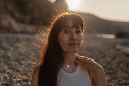 Woman, sunset, beach. Serene middle-aged woman enjoying golden hour light on a tranquil pebble shore, her hair illuminated.