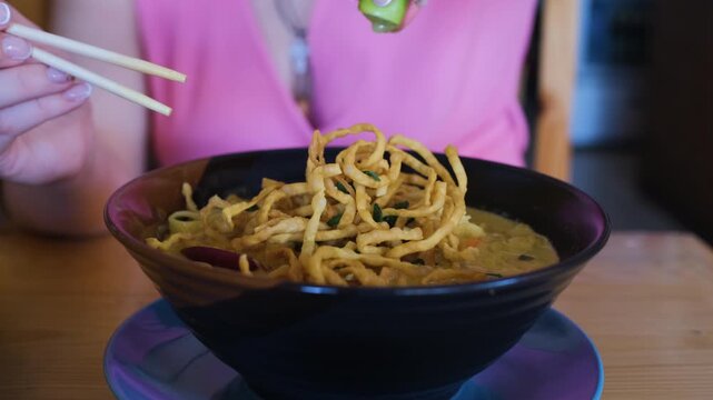 Close-up of a young woman squeezing fresh lime juice over a bowl of Khao Soi noodle curry before eating at a restaurant, slow motion. Thai food concept