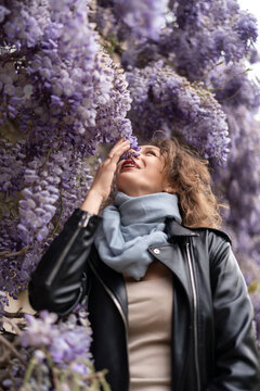 Spring garden beauty, smiling young woman smelling purple wisteria blossoms outdoors during seasonal bloom