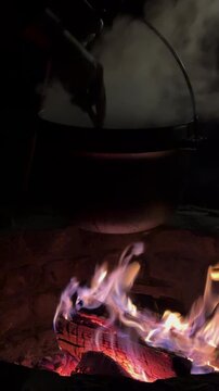 VERTICAL, CLOSE UP: Stirring steaming stew in a cast iron pot suspended above a glowing firepit. Flames, smoke and burning logs create warm, rustic cooking moment on autumn evening in the countryside