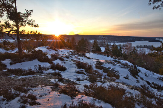 Golden sunrise bursting over a snowy winter forest in Dalsland, Sweden, with vibrant sun rays illuminating a frozen landscape and pine trees under a warm orange and yellow sky