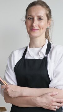 Confident female artist in a black apron holding brushes with her arms crossed, looking at the camera and smiling gently against a clean white background, showcasing her creative profession