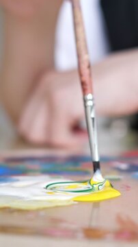 Talented artist's hand dipping a paintbrush into yellow and white paint on a palette, expertly mixing colors to create a new shade for a masterpiece in a creative studio workshop