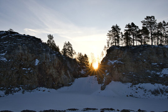 Impressive rock quarry in Sweden during winter with the rising sun peeking through cliffs and pine trees, casting long shadows across the snow-covered ground at Lake arr area.