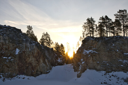 Impressive rock quarry in Sweden during winter with the rising sun peeking through cliffs and pine trees, casting long shadows across the snow-covered ground at Lake arr area.