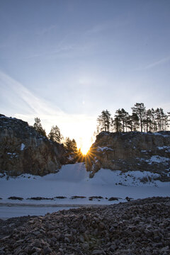 Impressive rock quarry in Sweden during winter with the rising sun peeking through cliffs and pine trees, casting long shadows across the snow-covered ground at Lake arr area.