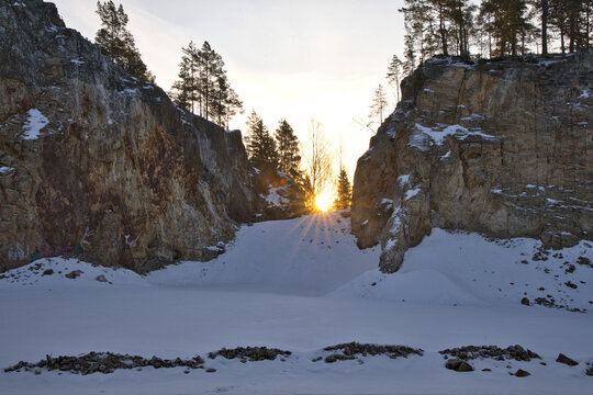 Impressive rock quarry in Sweden during winter with the rising sun peeking through cliffs and pine trees, casting long shadows across the snow-covered ground at Lake arr area.