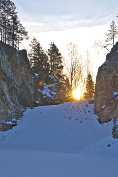 Impressive rock quarry in Sweden during winter with the rising sun peeking through cliffs and pine trees, casting long shadows across the snow-covered ground at Lake arr area.