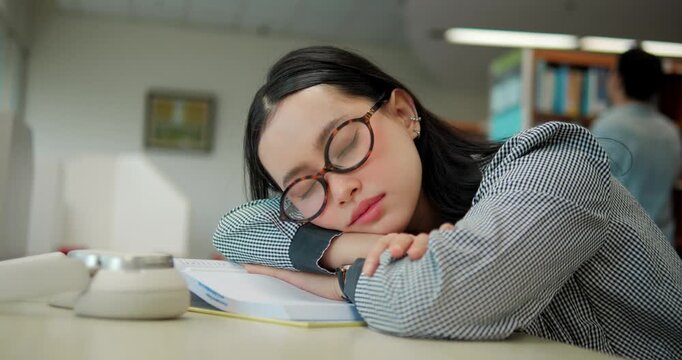 Girl rests eyes at desk, glasses askew. She sleeps peacefully in the library, head on open book.