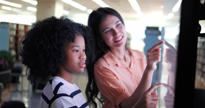 Young student girl and teacher explore interactive screen together. Girl reaches upward, curious and engaged. Woman smiles, guiding her attentively