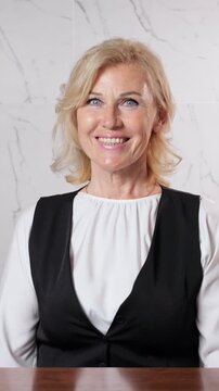 Portrait of a confident and charming middle aged businesswoman sitting at a desk, looking directly at the camera with a friendly and welcoming smile, expressing positivity and success